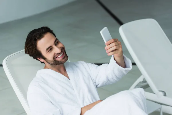 Hombre sonriente en albornoz tomando selfie en el teléfono inteligente en el centro de spa - foto de stock