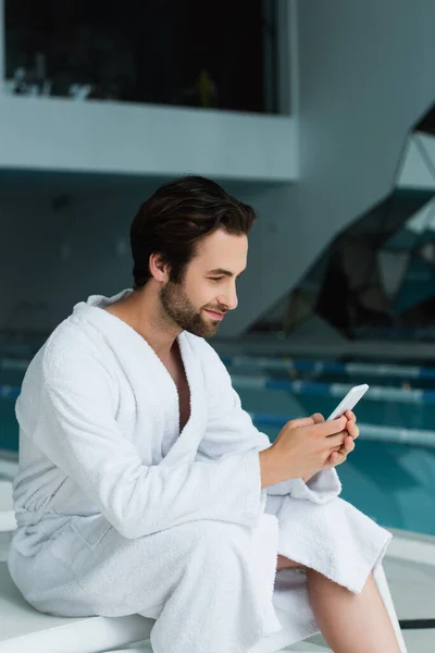 Homem de roupão de banho usando telefone celular na cadeira deck no centro de spa — Fotografia de Stock