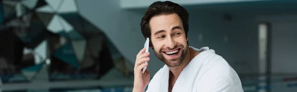 Улыбающийся мужчина в халате разговаривает по смартфону в спа-центре, баннер — стоковое фото