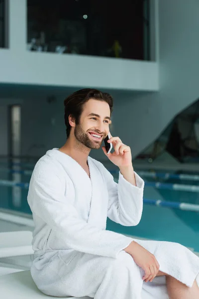 Cheerful man talking on smartphone near blurred swimming pool in spa center — Stock Photo