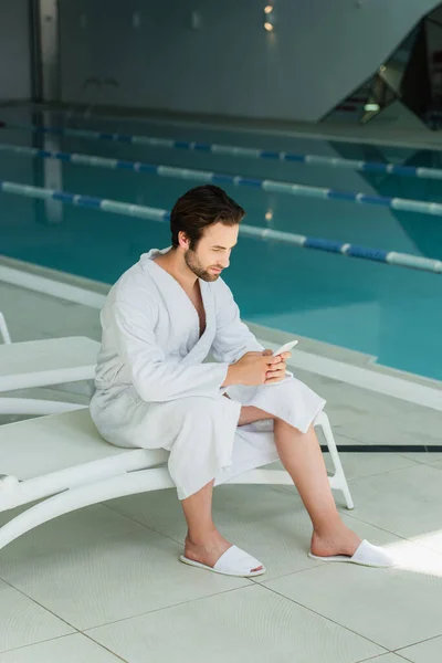 Hombre joven usando el teléfono celular en la silla de cubierta cerca de la piscina en el centro de spa - foto de stock