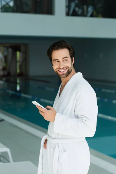 Homem positivo em roupão segurando smartphone no centro de spa — Fotografia de Stock