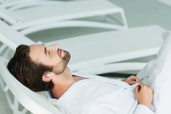 Homme en écouteur sans fil relaxant sur chaise longue dans le centre de spa — Photo de stock
