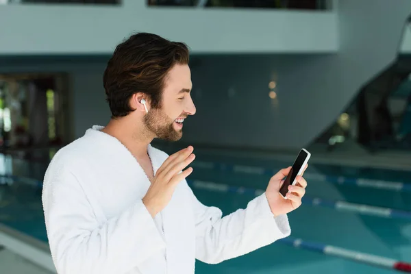 Side view of smiling man in earphone having video call on smartphone in spa center — Stock Photo