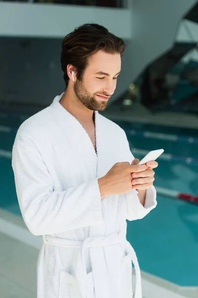 Young man in wireless earphone and bathrobe using smartphone in spa center — Stock Photo
