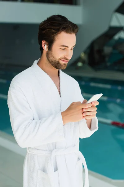 Hombre joven con auriculares inalámbricos y albornoz con teléfono inteligente en el centro de spa - foto de stock