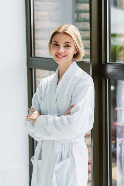 Smiling woman in bathrobe standing with crossed arms near window in spa center — Stock Photo