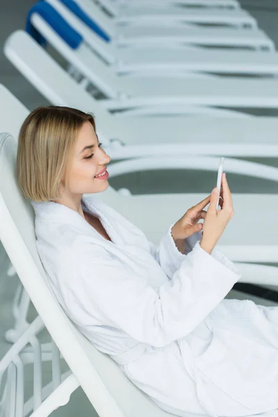 Mujer sonriente en albornoz blanco usando teléfono inteligente en silla de cubierta en el centro de spa - foto de stock