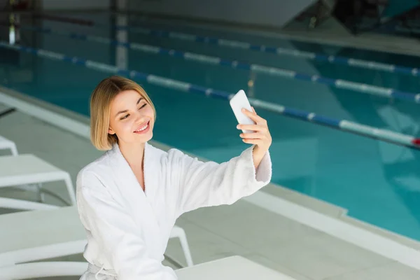 Donna sorridente scattare selfie su smartphone vicino alla piscina nel centro benessere — Foto stock