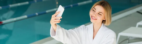 Mujer alegre en albornoz tomando selfie en el teléfono inteligente cerca de la piscina borrosa en el centro de spa, pancarta - foto de stock