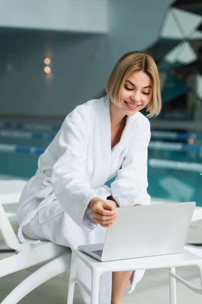 Fröhliche Frau im Bademantel mit Laptop auf Liegestuhl im Wellness-Center — Stockfoto