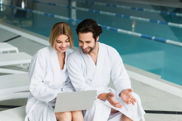 Pareja sonriente en albornoces usando portátil en la silla de cubierta en el centro de spa - foto de stock