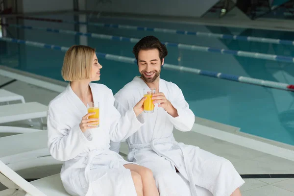 Smiling woman in bathrobe giving orange juice to boyfriend in spa center — Stock Photo