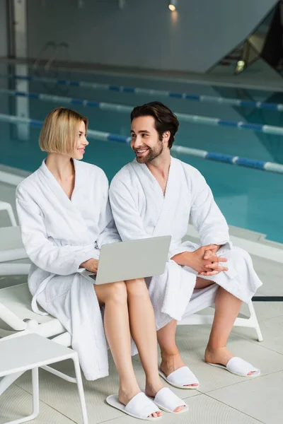 Junges Paar in Bademänteln lächelt einander an, während es im Wellness-Center Laptop benutzt — Stockfoto