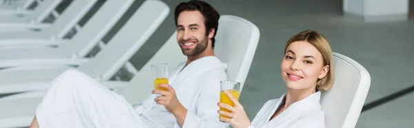 Mujer sonriente en albornoz sosteniendo jugo de naranja cerca de novio borroso en el centro de spa, pancarta - foto de stock