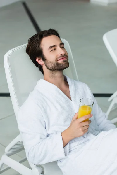 Jovem de roupão branco segurando suco de laranja enquanto relaxa no centro de spa — Fotografia de Stock