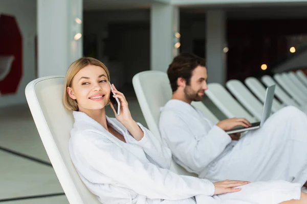 Positive woman in bathrobe talking on smartphone near blurred boyfriend with laptop in spa center — Stock Photo