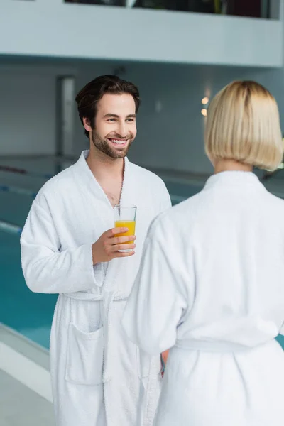 Homem sorridente em roupão de banho branco segurando vidro de suco de laranja perto de namorada borrada e piscina no centro de spa — Fotografia de Stock