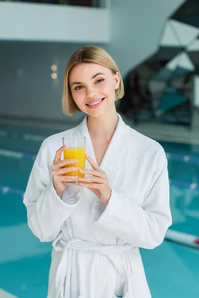 Young woman holding glass of orange juice near blurred swimming pool in spa center — Stock Photo