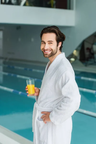 Young man in white bathrobe holding glass of orange juice near blurred swimming pool in spa center — Stock Photo