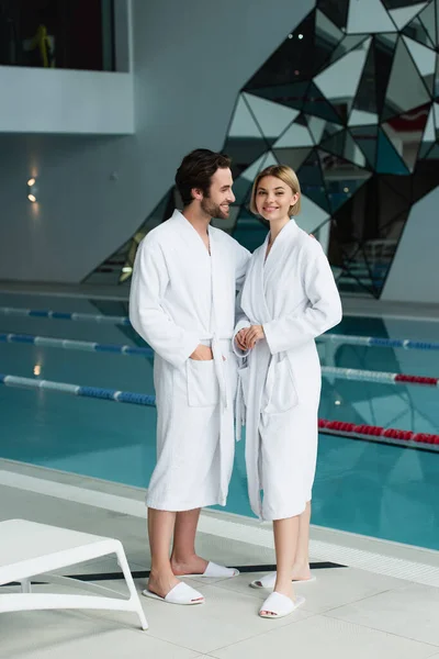 Pareja sonriente en albornoces de pie cerca de la piscina en el centro de spa - foto de stock