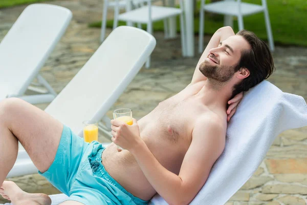Hombre sonriente en baúles de natación sosteniendo un vaso de jugo de naranja en la silla de cubierta en el resort - foto de stock