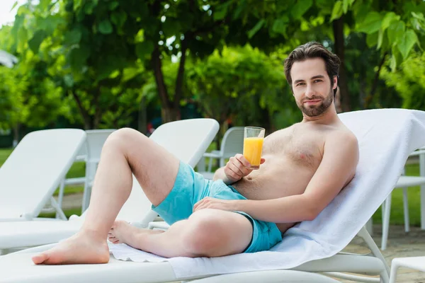 Young man in swimming trunks holding orange juice on deck chair on resort — Stock Photo