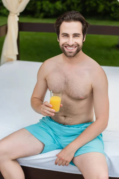 Hombre sonriente en bañador con zumo de naranja en la cama del salón en el resort - foto de stock