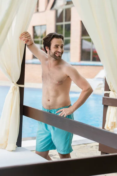 Hombre positivo en bañadores tomados de la mano en la cadera cerca de la cama del salón y la piscina borrosa - foto de stock