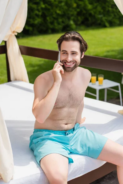 Homme souriant dans les maillots de bain parler sur smartphone sur lit lounge à l'extérieur — Photo de stock