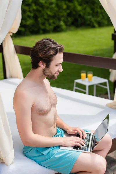Side view of man in swimming trunks using laptop on lounge bed outdoors — Stock Photo