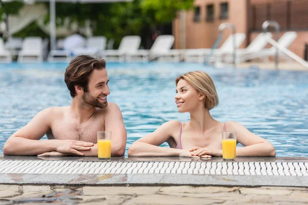 Cheerful woman in swimming pool looking at boyfriend near orange juice on poolside — Stock Photo