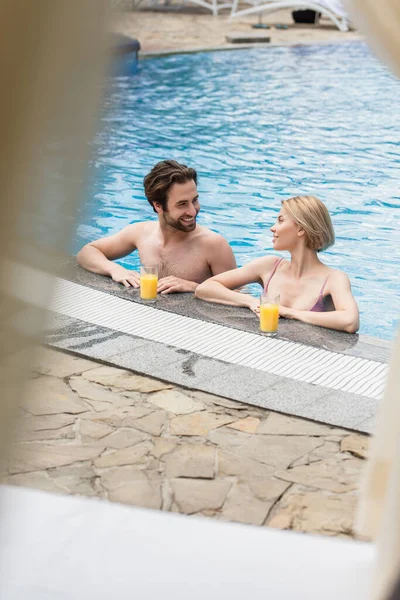 Lächelnder Mann schaut Freundin im Schwimmbad in der Nähe von Orangensaft-Gläsern am Pool an — Stockfoto