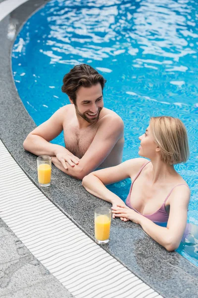 Вид на позитивную пару, беседующую в бассейне рядом с апельсиновым соком на берегу бассейна — стоковое фото