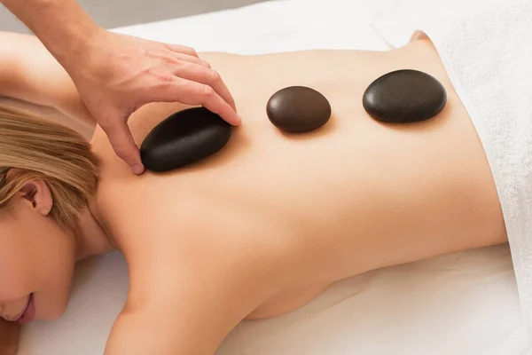 Vue de dessus du masseur mettant la pierre de spa sur le dos de la jeune femme sur la table de massage — Photo de stock