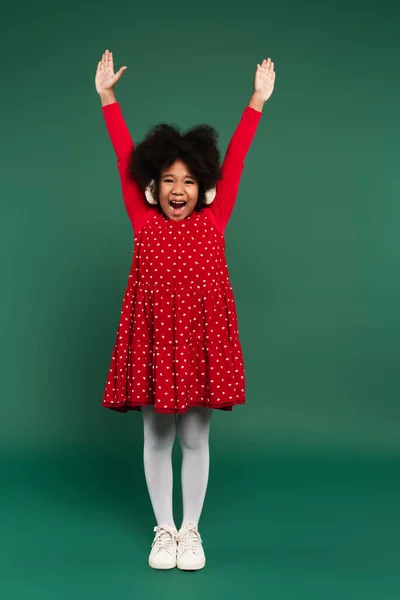 Alegre niño afroamericano en orejeras y vestido rojo levantando las manos sobre fondo verde - foto de stock