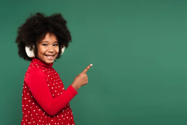 Smiling african american kid in ear muffs pointing with finger isolated on green — Stock Photo