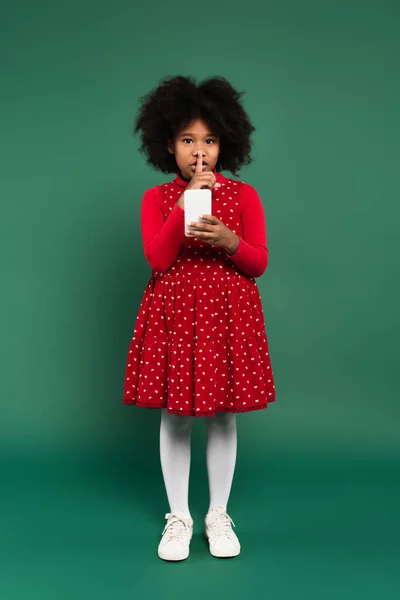 Niño afroamericano en vestido rojo mostrando gesto secreto y usando teléfono inteligente sobre fondo verde - foto de stock