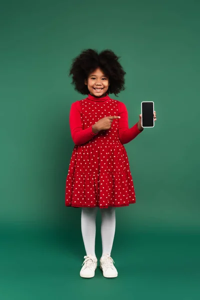 Lächelnde Afroamerikanerin in Kleid zeigt auf Smartphone mit leerem Bildschirm auf grünem Hintergrund — Stockfoto