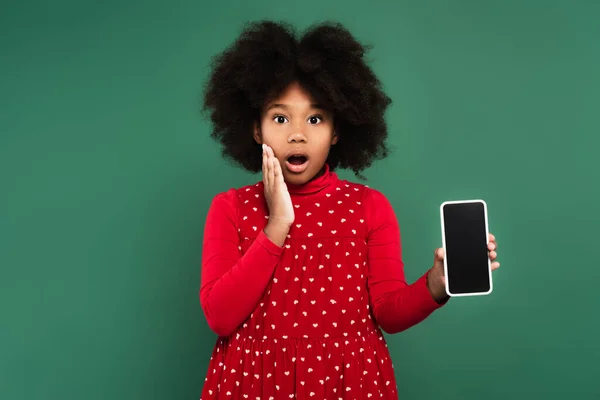 Niño afroamericano sorprendido en vestido rojo sosteniendo teléfono celular con pantalla en blanco aislado en verde - foto de stock