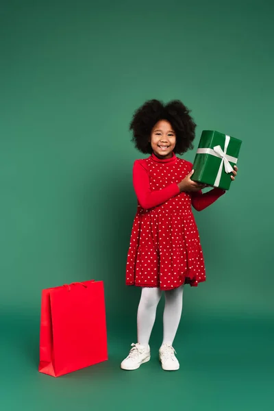 Glückliche afrikanisch-amerikanische Kind hält Geschenkbox mit Band in der Nähe Einkaufstasche auf grünem Hintergrund — Stockfoto