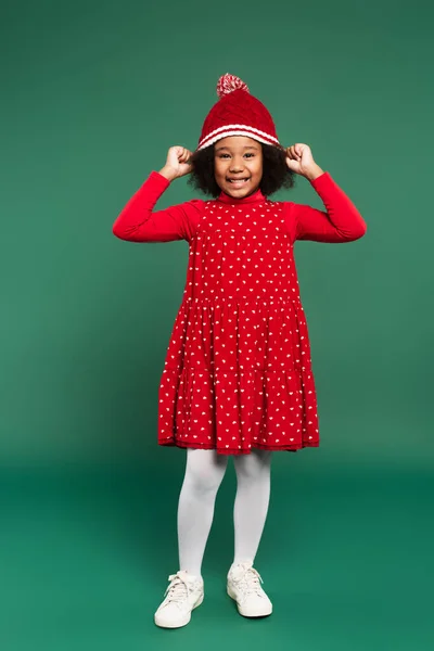Piena lunghezza di sorridente ragazza afroamericana in abito rosso indossando cappello su sfondo verde — Foto stock