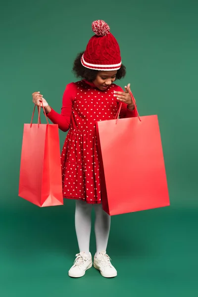Pleine longueur de gamin afro-américain en robe et chapeau tricoté regardant sac à provisions rouge sur fond vert — Photo de stock