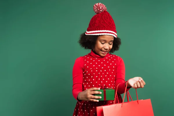 Enfant afro-américain dans un chapeau tenant un sac à provisions et une boîte cadeau isolés sur vert — Photo de stock