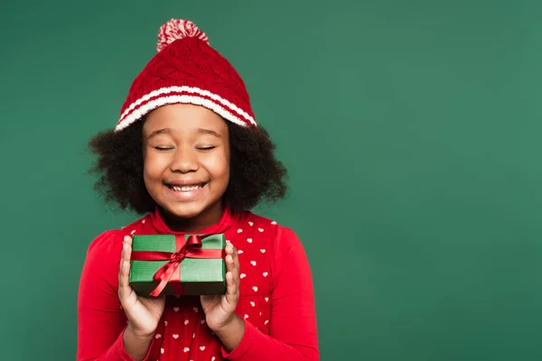 Joyful african american kid in warm hat closing eyes while holding gift box isolated on green — Stock Photo