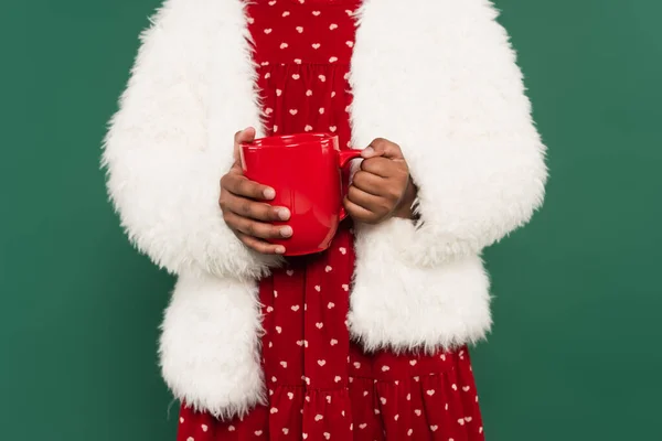 Vista recortada de chica afroamericana en chaqueta esponjosa sosteniendo taza aislada en verde - foto de stock