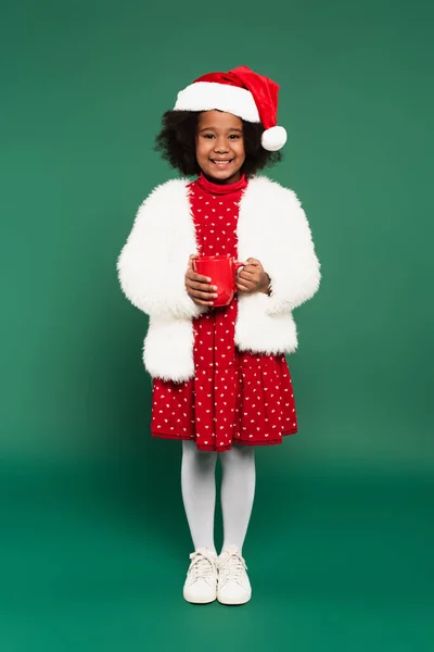 African american child in santa hat and fluffy jacket holding cup on green background — Stock Photo