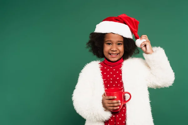 Sonriente niño afroamericano en sombrero de santa y chaqueta esponjosa sosteniendo taza roja aislada en verde - foto de stock