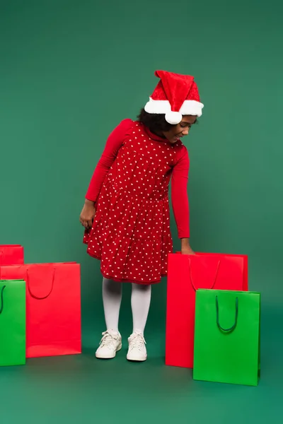 Вид сбоку позитивной африканской девушки в шляпе санта и красном платье, смотрящей на сумку с покупками на зеленом фоне — стоковое фото