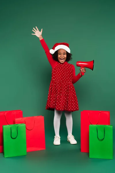 Positive afrikanisch-amerikanische Mädchen mit Weihnachtsmütze hält Lautsprecher in der Nähe von Einkaufstüten auf grünem Hintergrund — Stockfoto